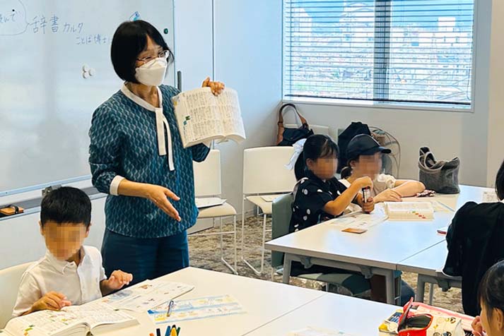 教室の写真。大きな机がふたつ、それぞれ4人の子どもが座っており、辞書を見せながら説明する柏野先生