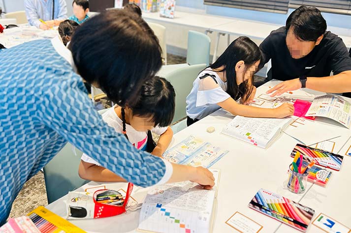 辞書で調べている親子と、女の子に調べ方のヒントを教えている柏野先生の写真